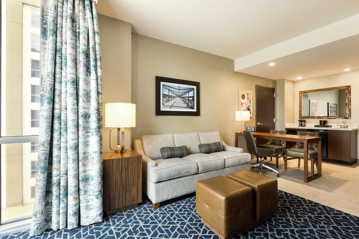 A hotel room with a sofa, ottoman, dining table, and a kitchen area.