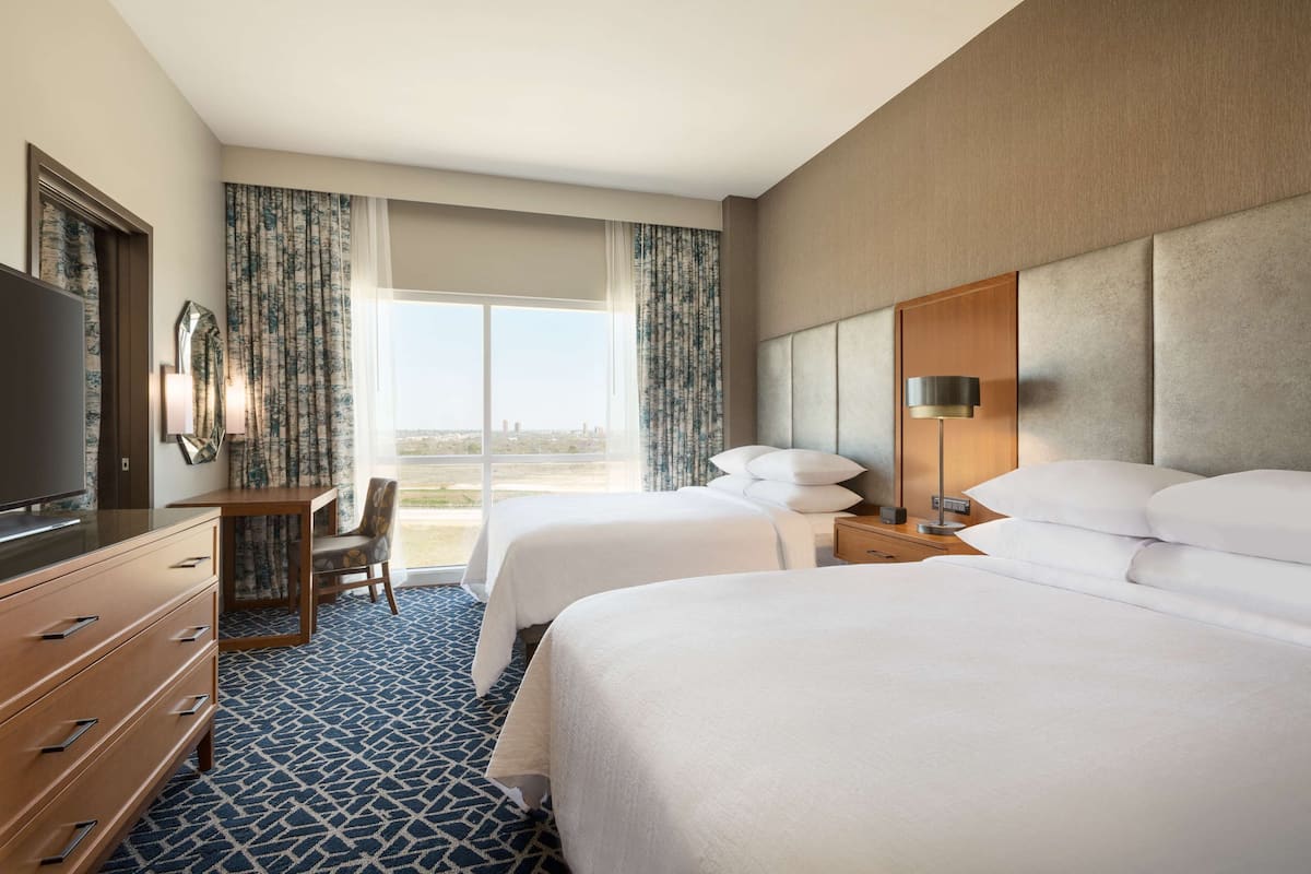A hotel room with two beds, a desk, a TV, and a large window with curtains.