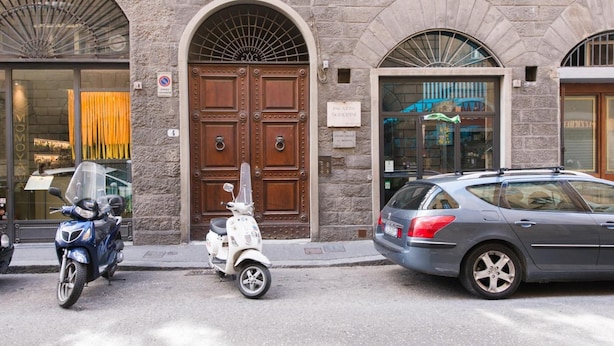 A street view with a stone building featuring a large wooden door, a parked scooter, and a car. - Paravia Suite (Florence)