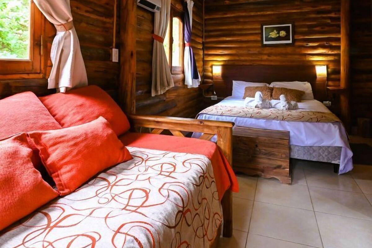 A cozy log cabin room with two beds, a wooden ceiling, and a window with curtains.