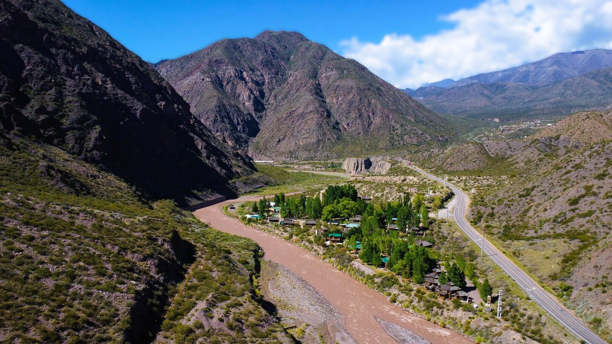 A mountainous landscape with a winding road, a small settlement with houses and greenery, and distant mountains.