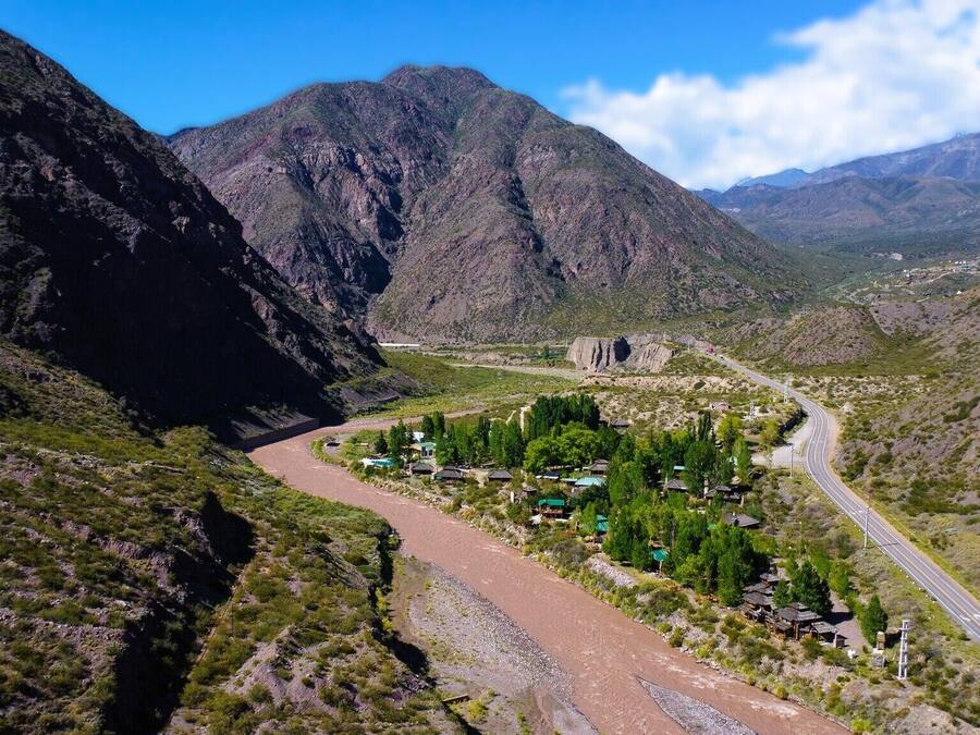A mountainous landscape with a winding road, a small settlement with houses and greenery, and distant mountains.