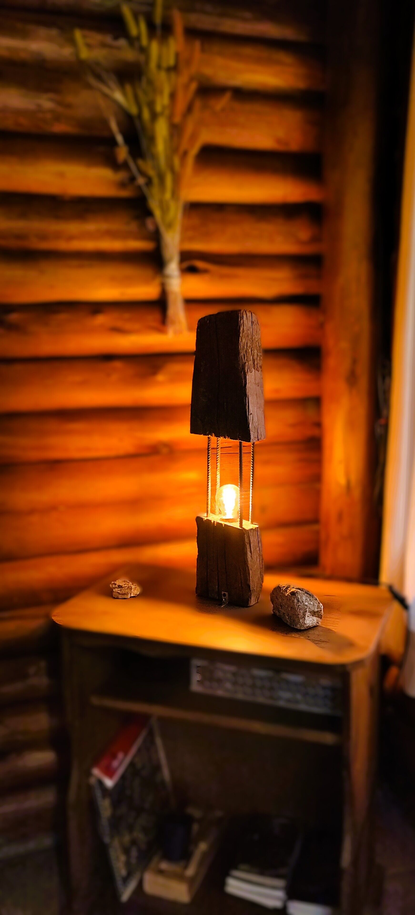 Interior rústico de una cabaña de madera, con una mesa central de madera que sostiene un farol, rodeada de libros y un pequeño adorno.