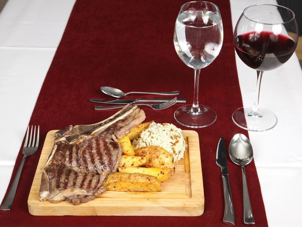 A wooden cutting board with grilled steak, roasted potatoes, and rice, accompanied by two wine glasses on a red tablecloth.