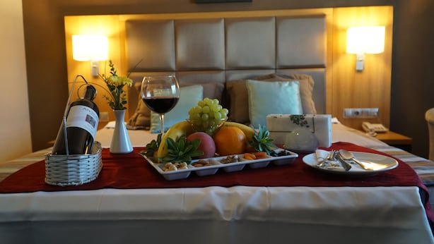 A neatly arranged breakfast tray with fruits, nuts, and a bottle of wine on a bed with a red blanket. - Efeler Hotel (Aydin)