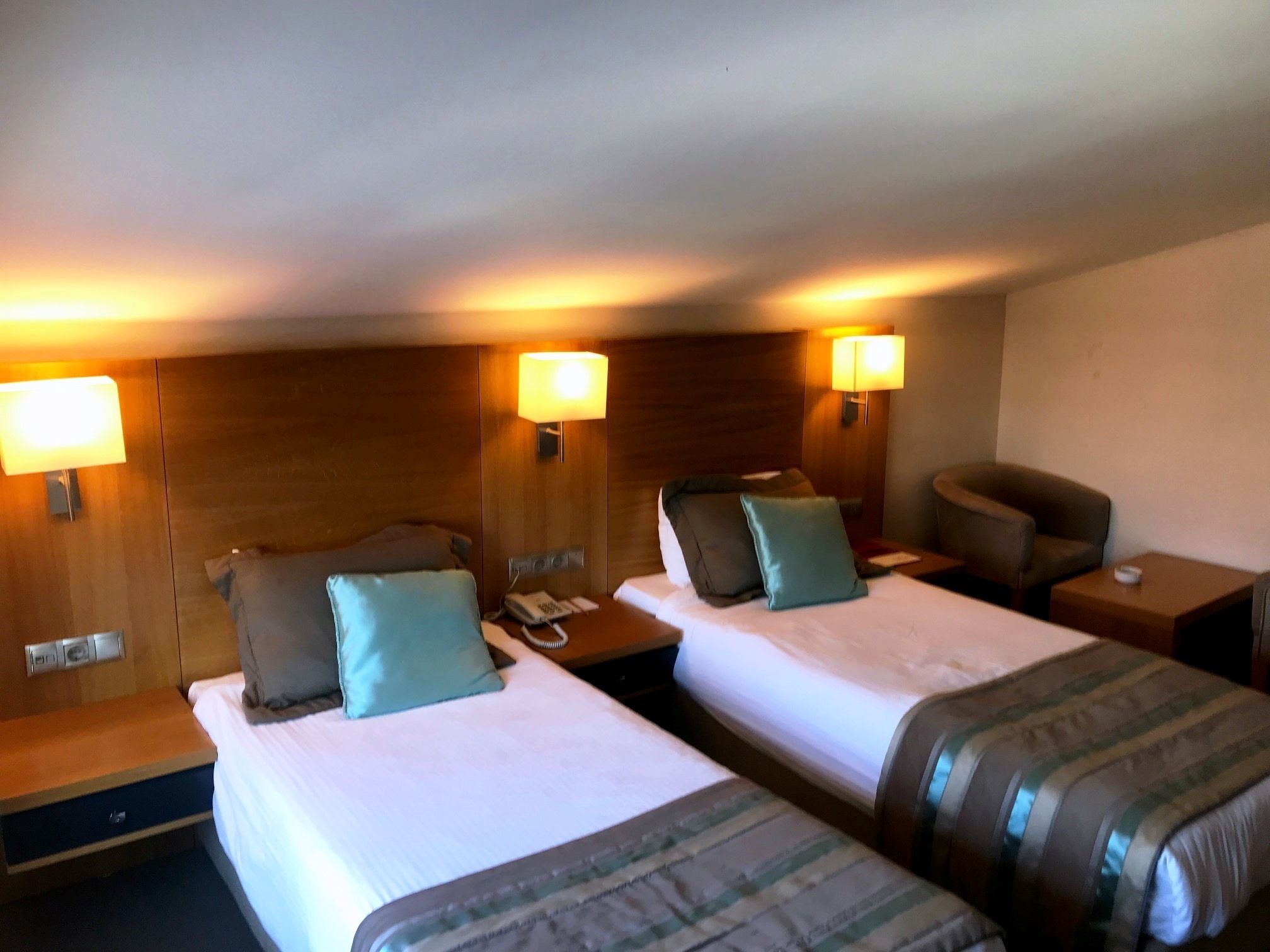 Habitación de hotel con dos camas, cada una con un cojín y una lámpara de noche. Hay una silla y una mesita con un teléfono encima.