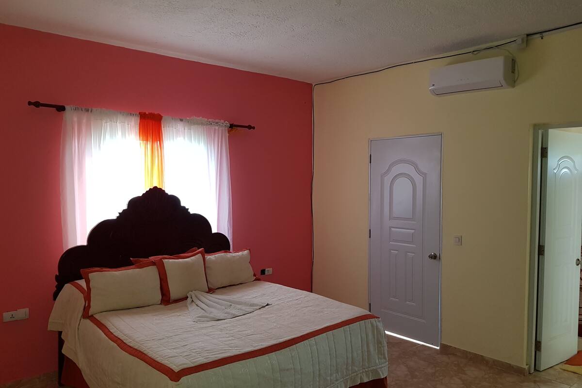 A bedroom with a bed, a window with curtains, a door, and an air conditioning unit.