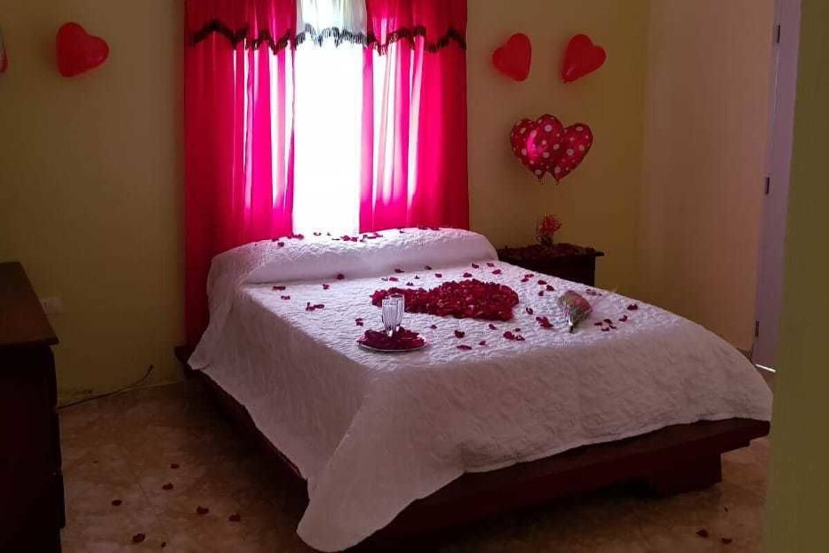 A bedroom with a bed covered in white linens, a bedside table with a vase, and a window with red curtains.