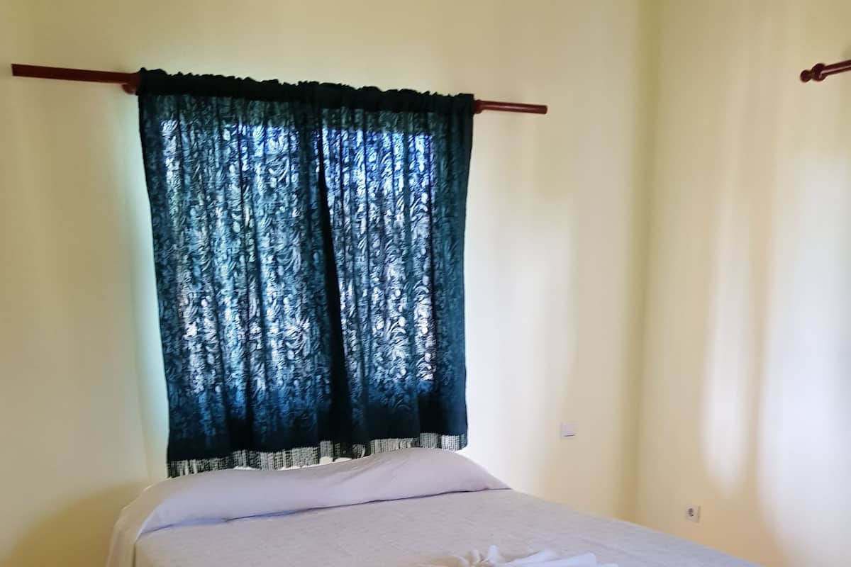 A single bed with a white bedspread and a folded towel on it, a dark curtain, and a tiled floor.