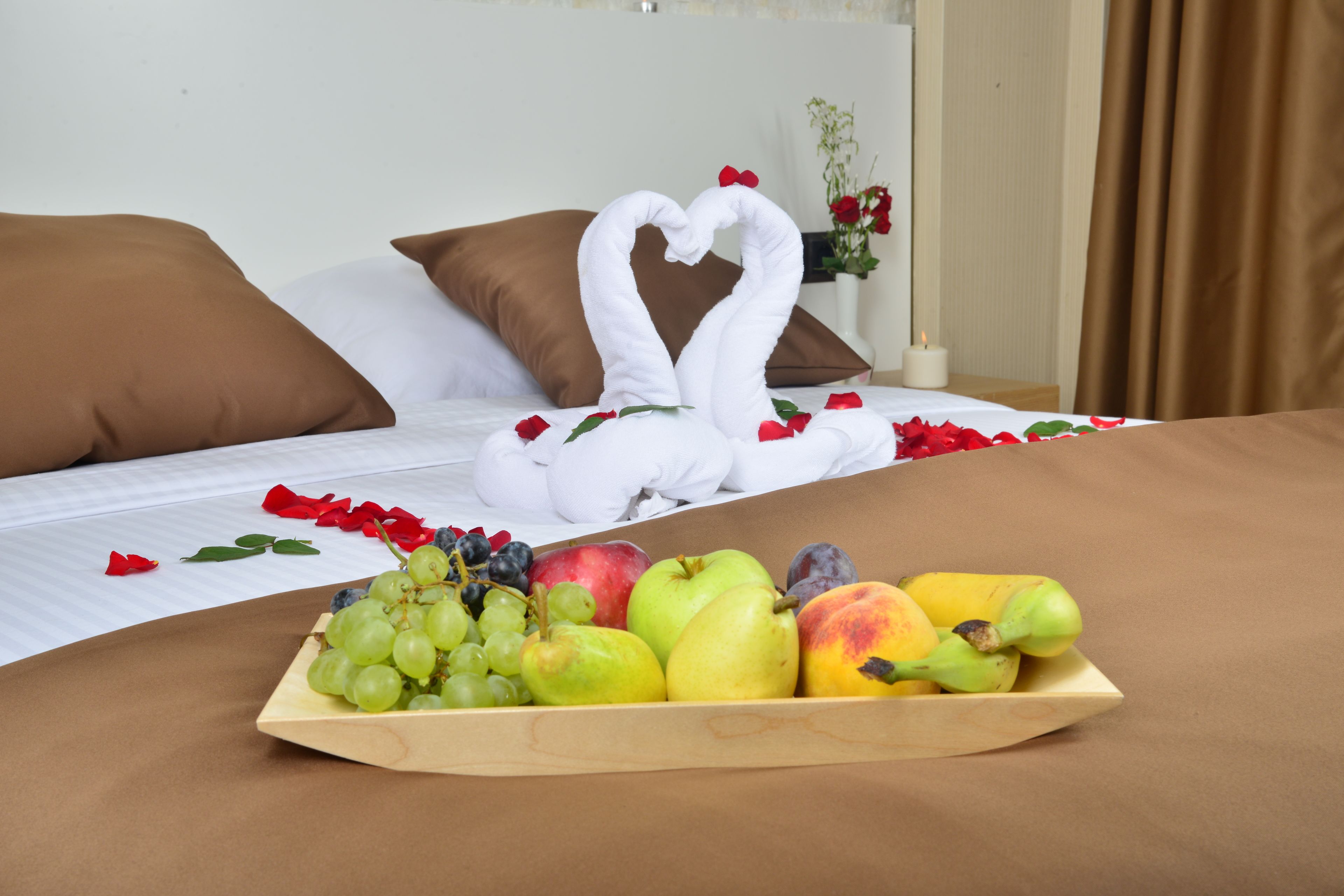 Una cama bien hecha con una toalla dispuesta en forma de corazón, un frutero de madera con uvas, manzanas y melocotones, y un florero con flores rojas a un lado.