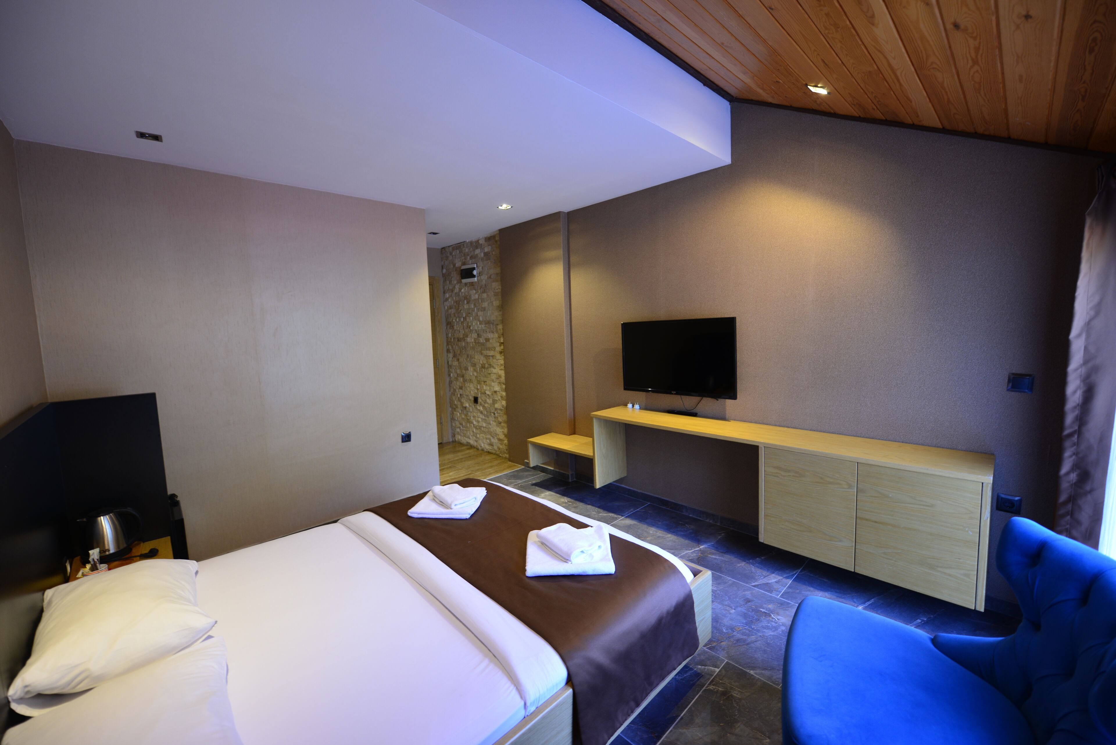A hotel room with a bed, a TV mounted on the wall, a seating area, and a stone wall.