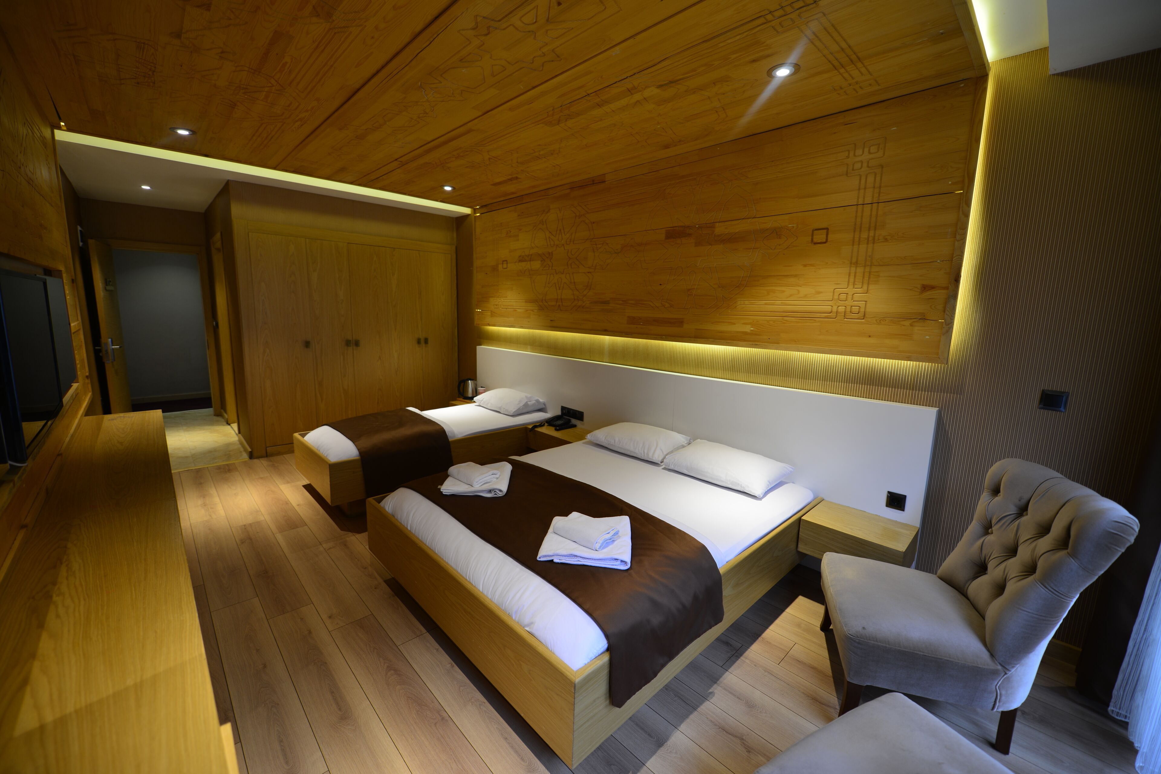 A modern bedroom with a wooden ceiling, a large bed with a headboard, a seating area with a chair, and a wardrobe.