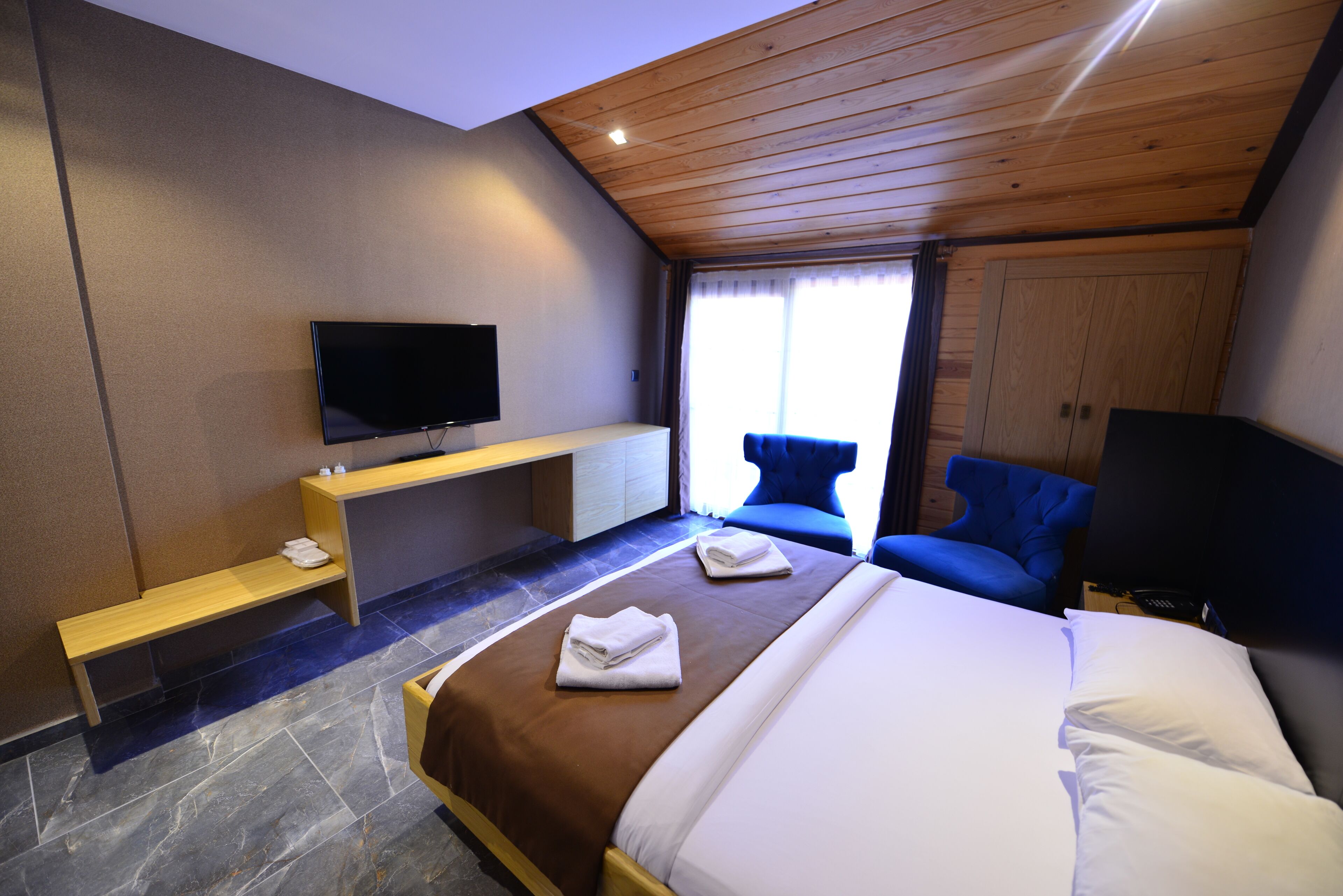A modern hotel room with a large bed, a flat-screen TV mounted on the wall, a wooden ceiling, and a seating area with a blue chair.