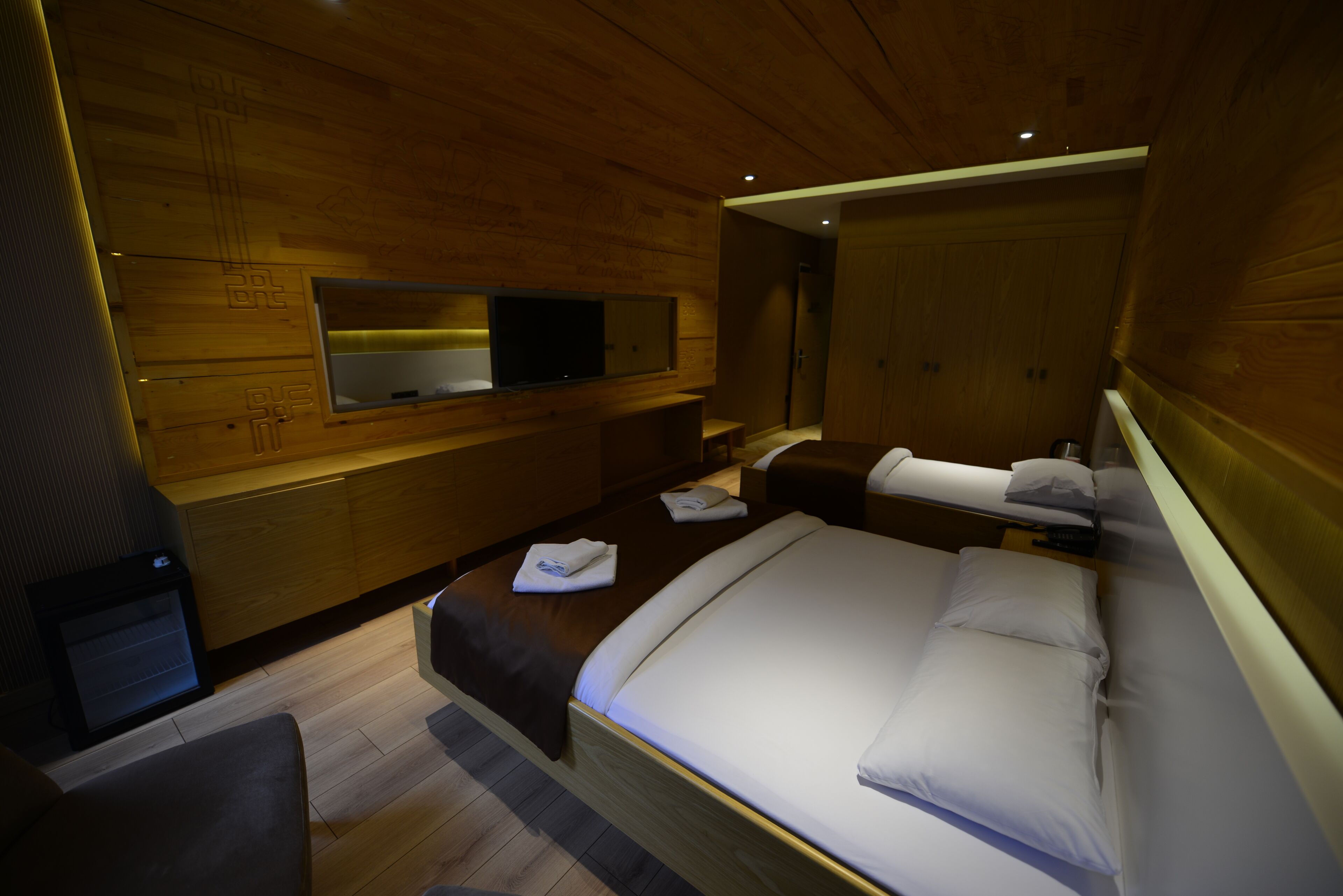 A modern hotel room with a wooden interior, a large bed, a flat-screen TV, and a seating area with a bench.