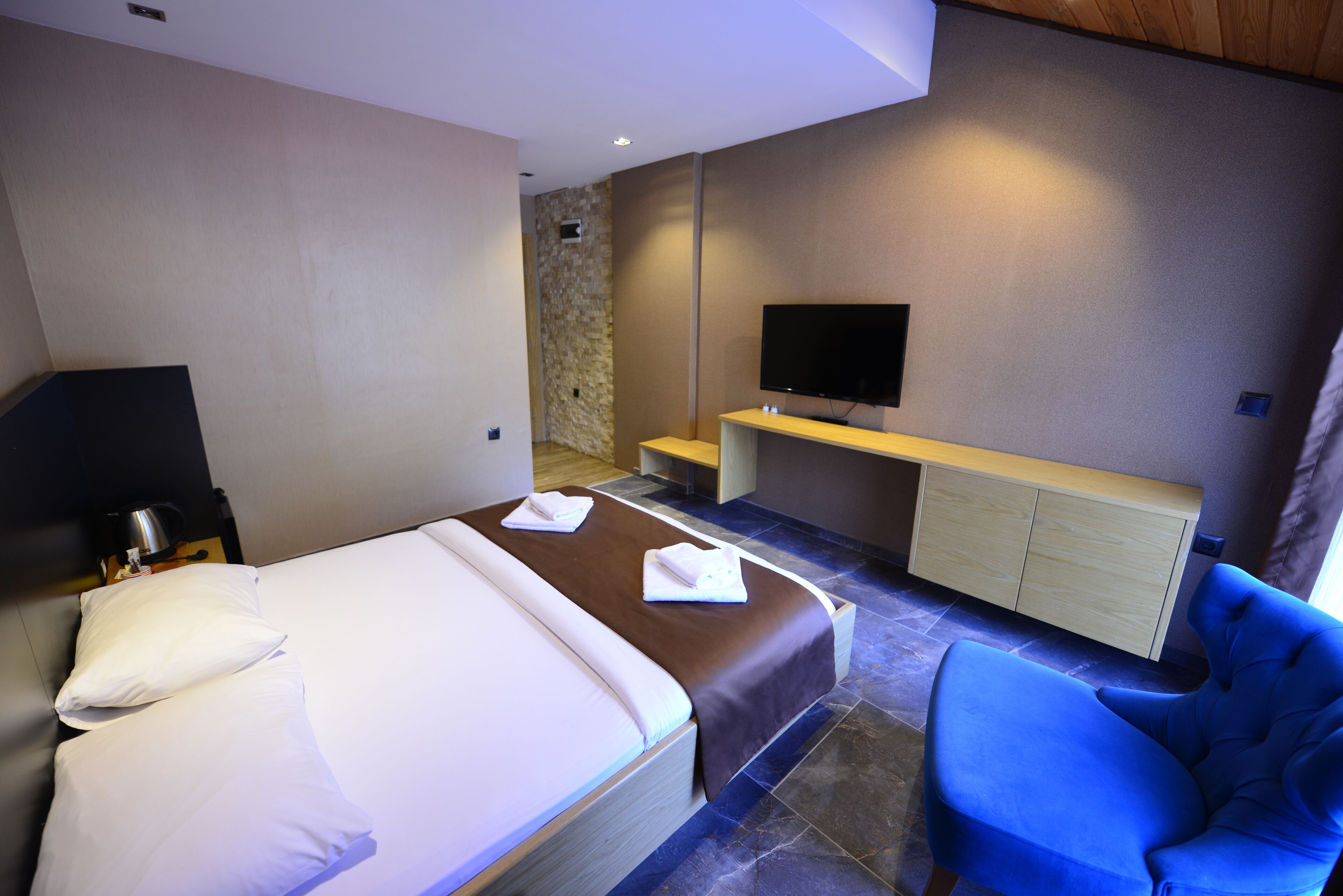 A modern hotel room with a large bed, a blue armchair, a flat-screen TV mounted on the wall, and a small wooden table.