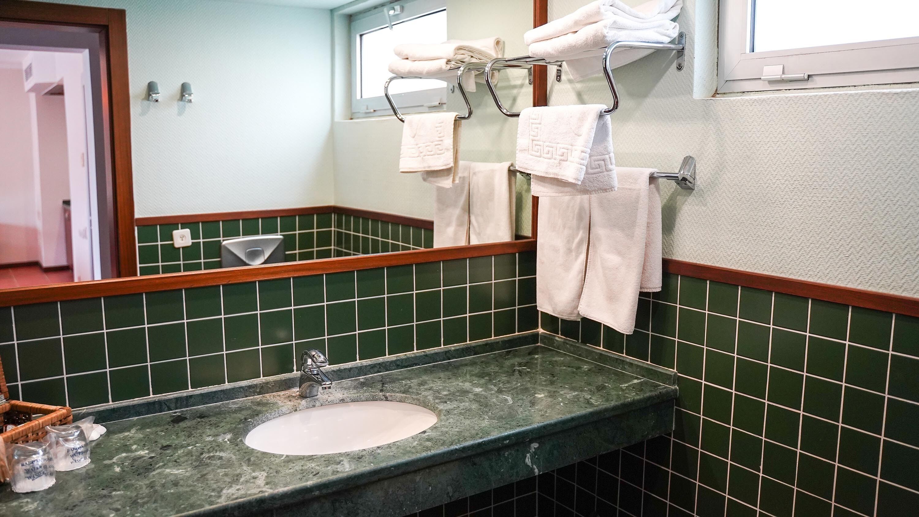 Un baño con pared de azulejos verdes, un lavabo de mármol y un perchero con toallas dobladas.
