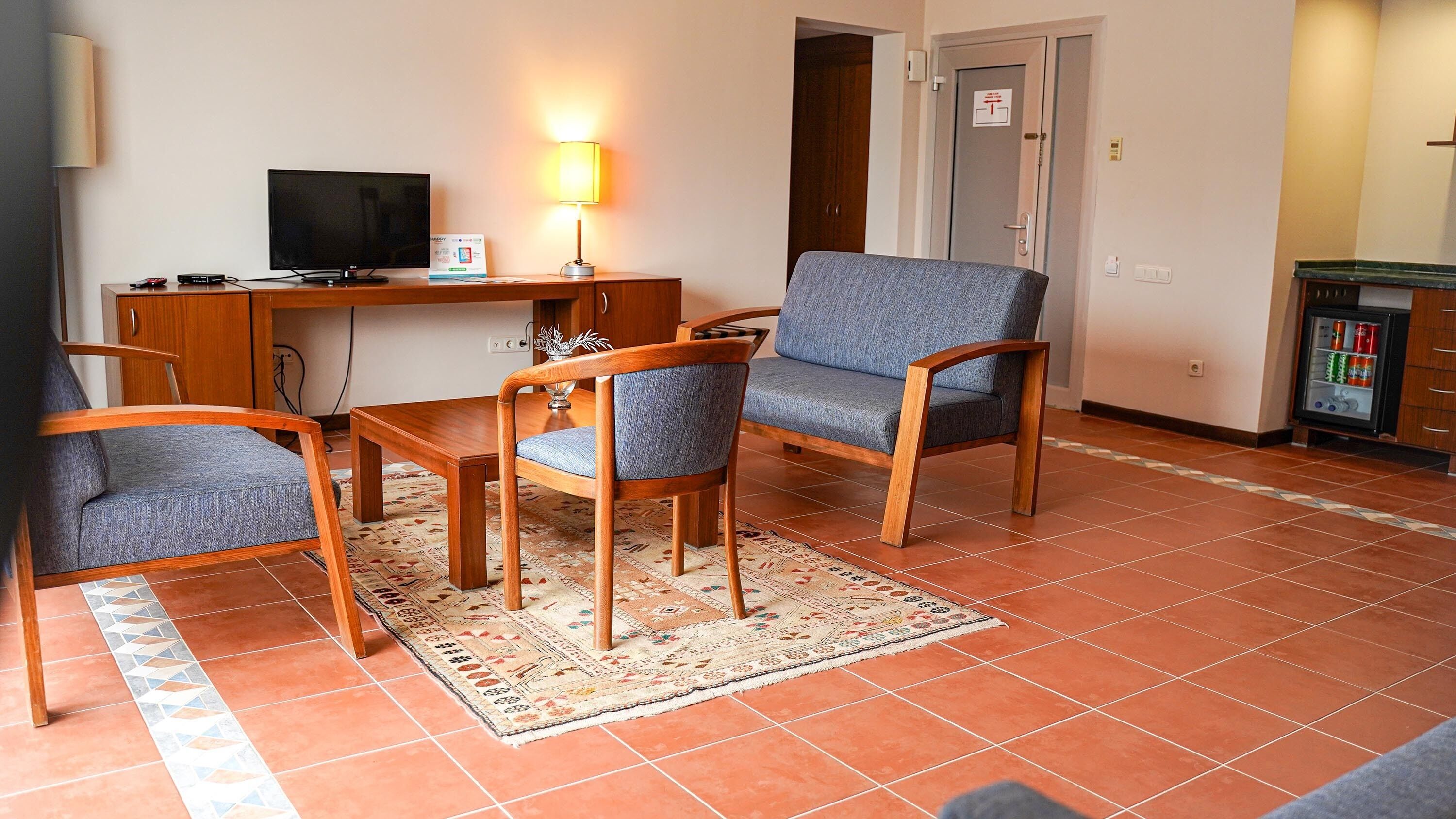 Quarto com escrivaninha de madeira, televisão, luminária e área de estar com cadeiras e tapete.