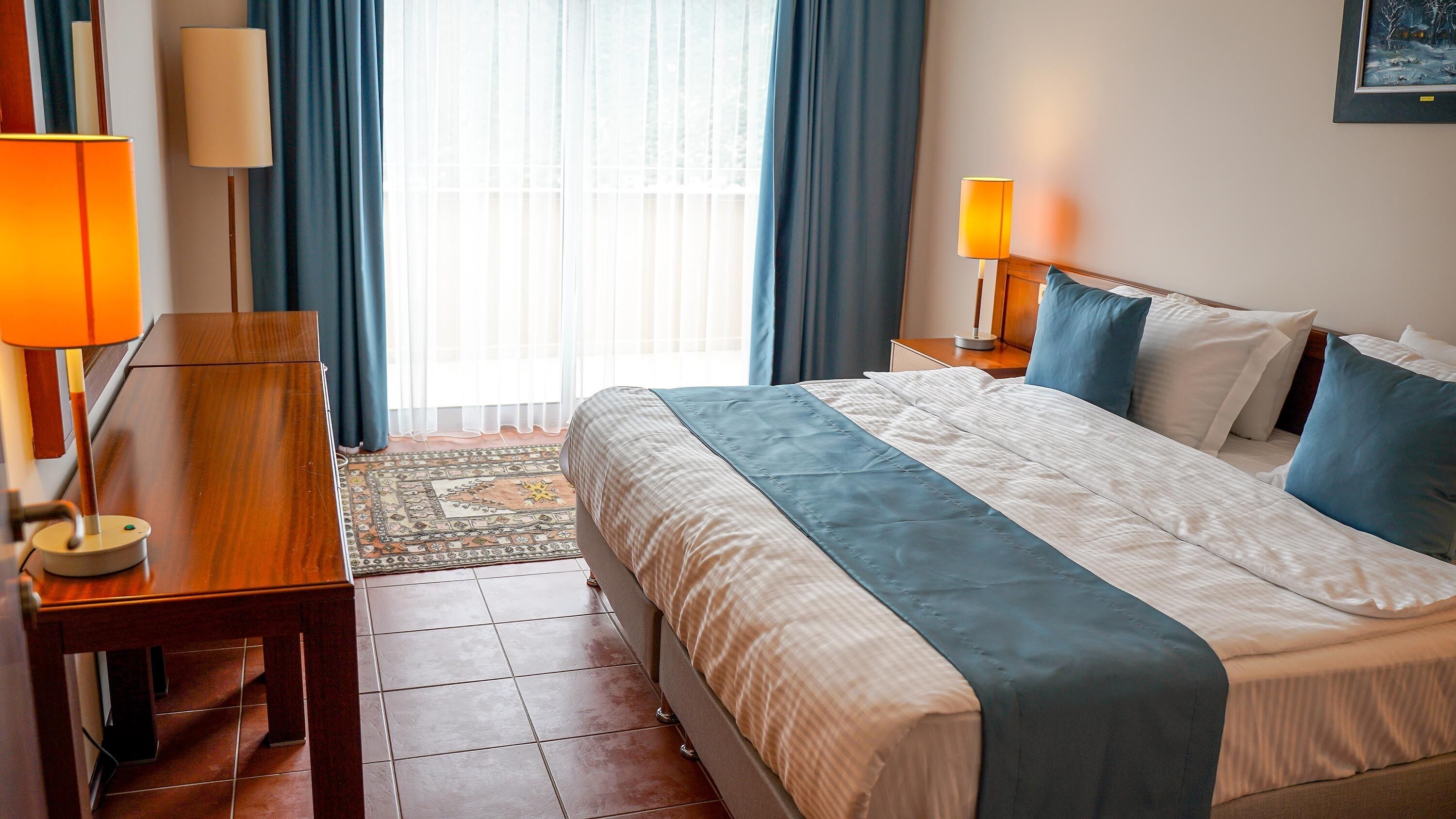 A hotel room with a bed, two bedside tables, a lamp, a rug, and a painting on the wall.