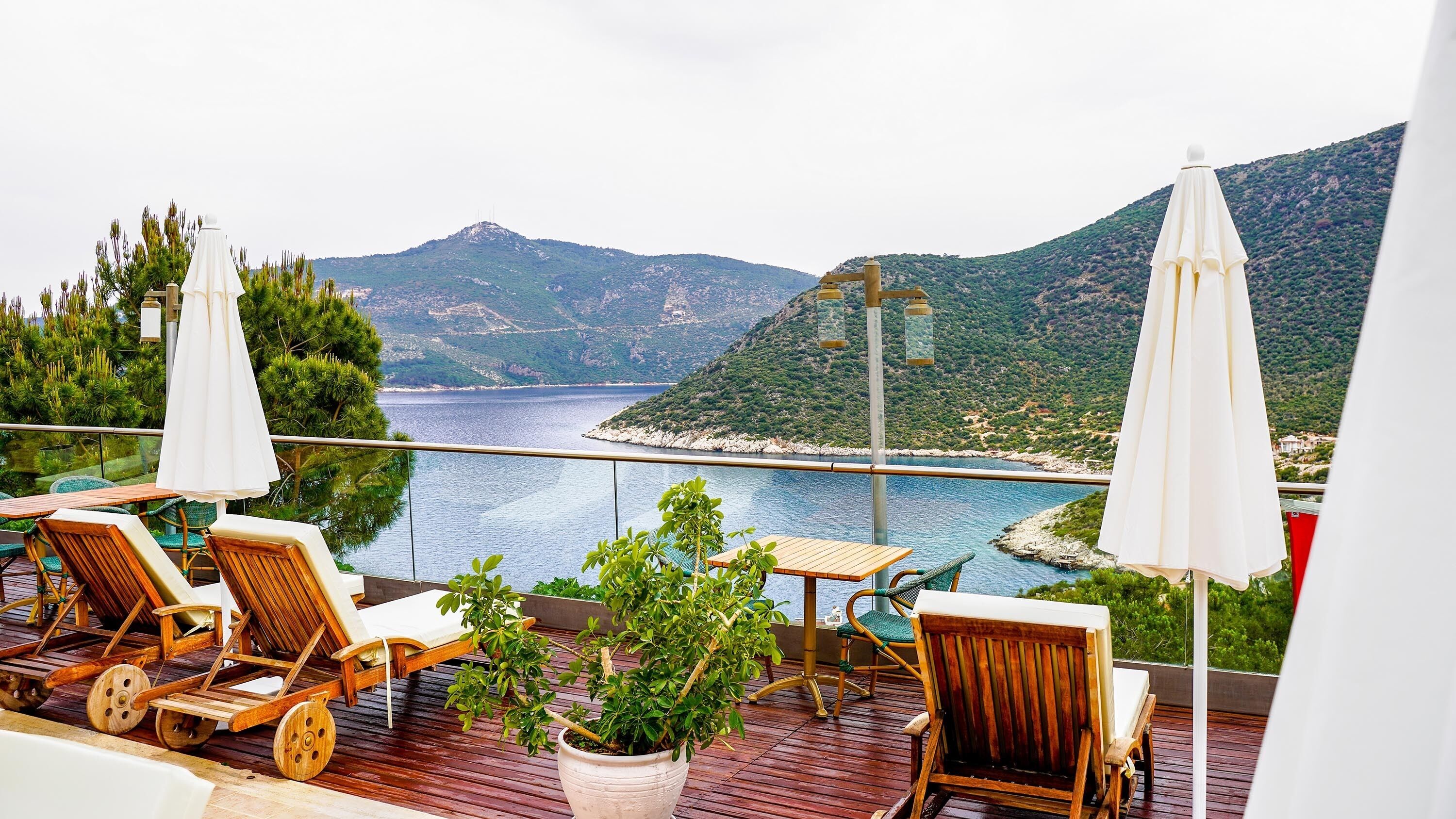 Una terraza con sillones de madera y una mesa, con vistas a un cuerpo de agua y un paisaje montañoso.