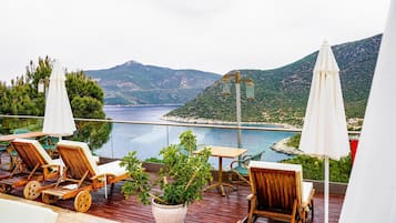 Una terraza con sillones de madera y una mesa, con vistas a un cuerpo de agua y un paisaje montañoso.