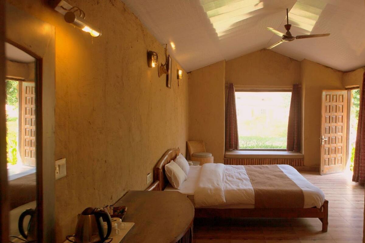 A bedroom with a bed, a desk with a kettle, a window with curtains, and a ceiling fan.