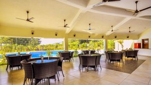 Un amplio espacio exterior con mesas y sillas, ventiladores de techo y vistas a una piscina y follaje.