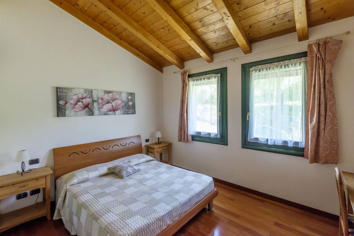 A bedroom with a wooden ceiling, a bed with a quilt, two windows with curtains, a nightstand with a lamp, and a wall-mounted painting.