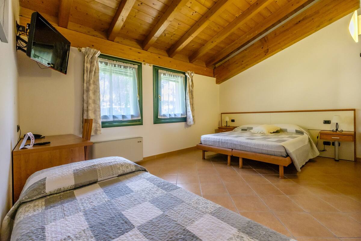 A bedroom with a wooden ceiling, two windows with curtains, a bed with a quilt, a bedside table, and a TV mounted on the wall.