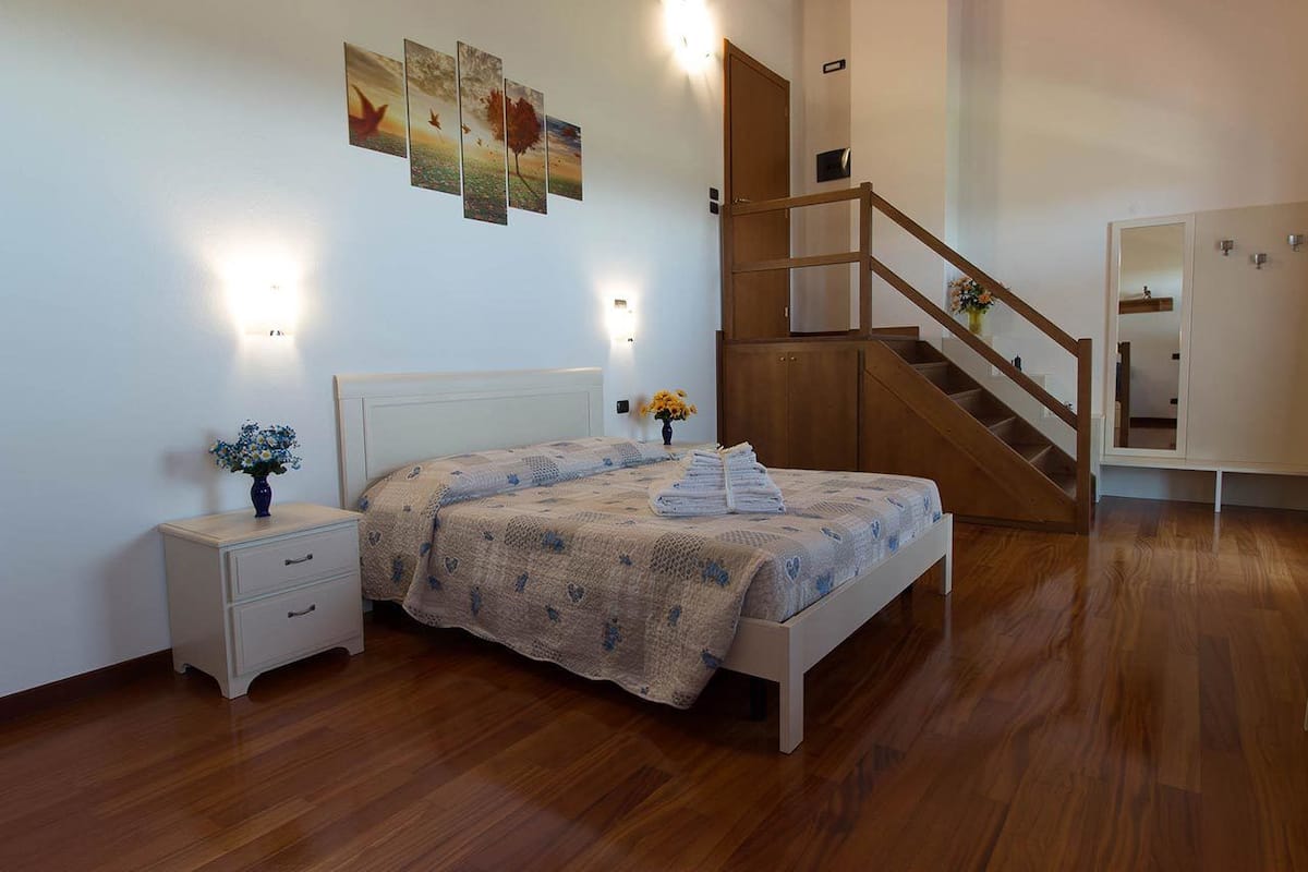 A bedroom with a wooden floor, a bed with a quilt, a nightstand with a vase, and a staircase.