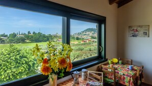 A room with a large window offering a view of a green landscape and distant buildings. There is a table with a colorful tablecloth and a vase of flowers on the windowsill. - Agriturismo Al Colle (Cavaion Veronese)