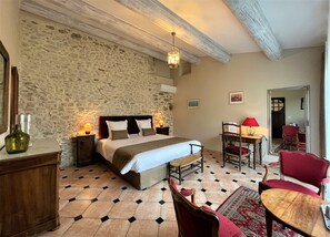 A bedroom with a stone wall, a bed with white bedding, a wooden nightstand, a red chair, a wooden table, and a mirror. - Le Mas Carré (Seguret)