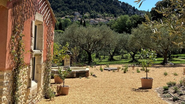 A courtyard with a building featuring a tiled roof and stone walls, surrounded by olive trees and a gravel path. - Le Mas Carré (Seguret)