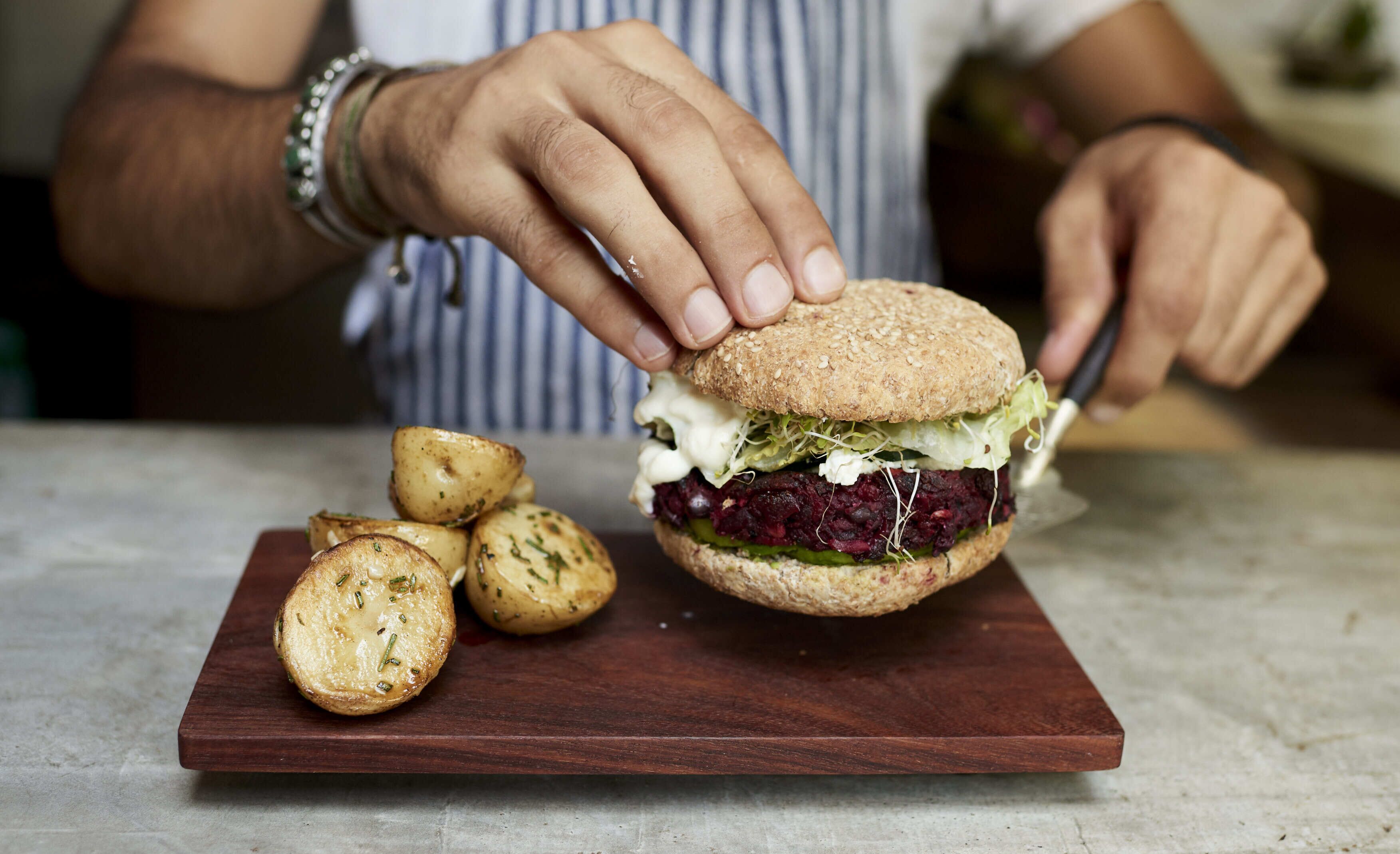 A person assembling a sandwich on a wooden board with a knife.
