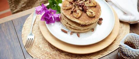 Une pile de crêpes sur une assiette, garnie de noix de pécan et de tranches de banane, accompagnée d’une tasse de café et d’un petit bol de miel.