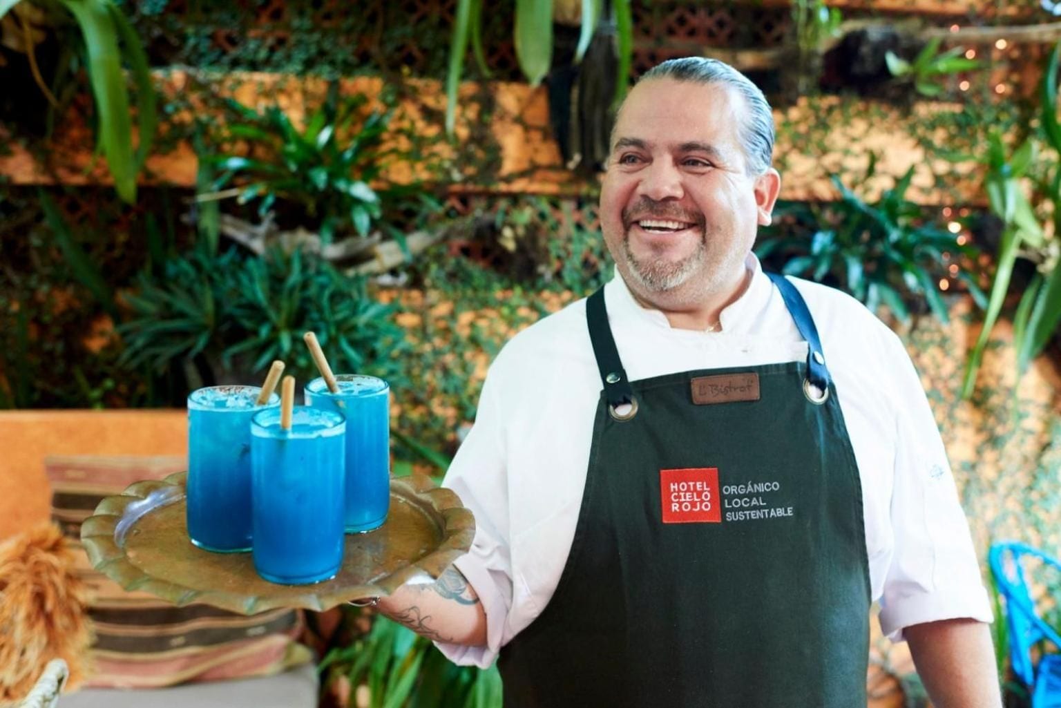 A person holding a tray with three blue drinks, wearing a black apron with a logo, in a setting with greenery and a patterned wall.