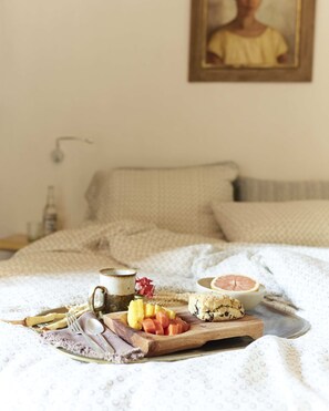 Un desayuno con una tabla de madera que contiene frutas, una galleta y una taza, sobre una cama con colcha y almohadas estampadas.