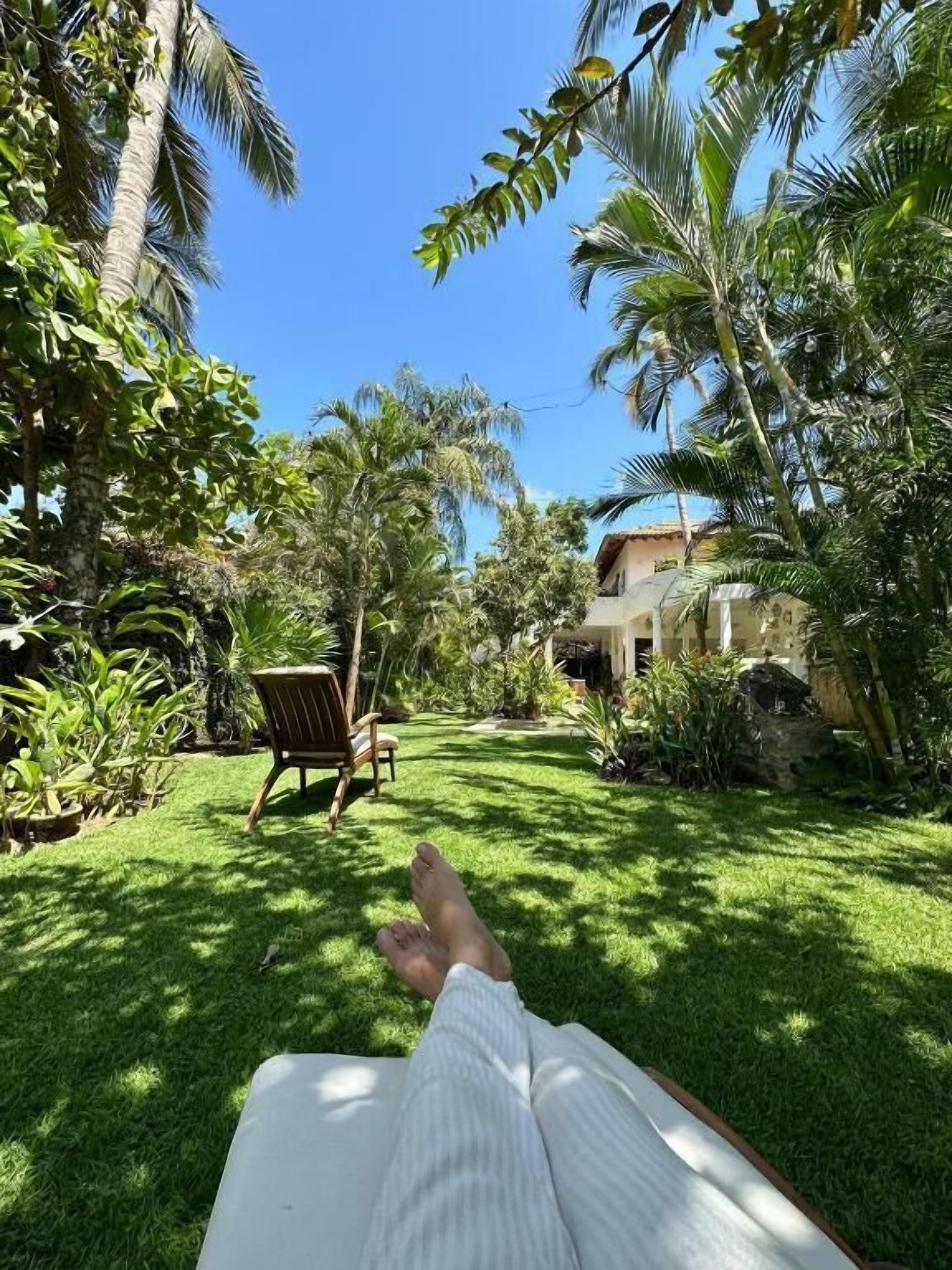 Una persona recostada en una silla de descanso en un área verde, con vista a un edificio y palmeras.