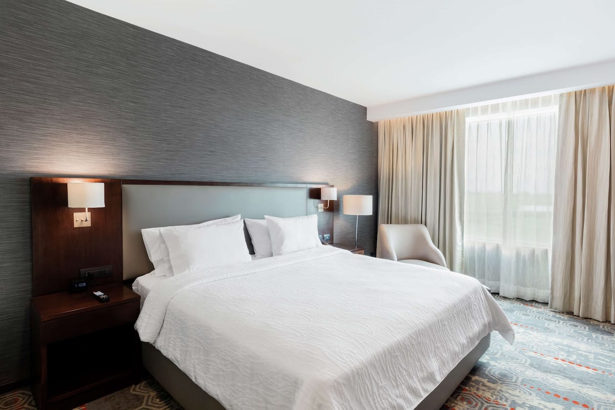 Una habitación de hotel con una cama grande, dos mesitas de noche con lámparas, una silla y un ventanal con cortinas.