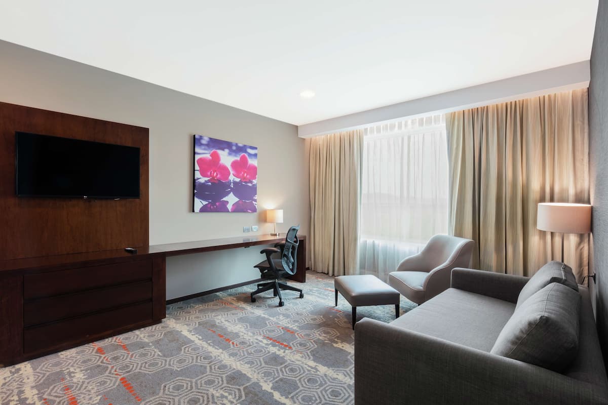 Habitación de hotel con televisión de pantalla plana montada en la pared, un escritorio con silla, un sofá y una ventana grande con cortinas.