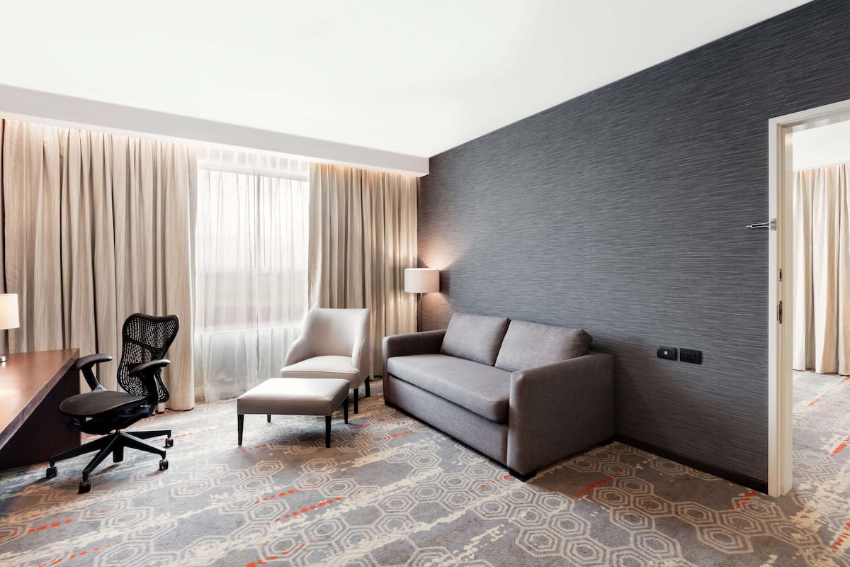Habitación de hotel con sofá gris, un sillón pequeño, un escritorio con lámpara y alfombra estampada.