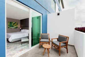 Een moderne hotelkamer met een bed, een klein tafeltje, twee stoelen en een glazen schuifdeur die uitkomt op een balkon met uitzicht op de lucht.