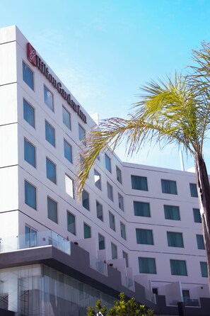 Een modern hotelgebouw met een opvallend logo op de gevel, meerdere ramen en een palmboom op de voorgrond.