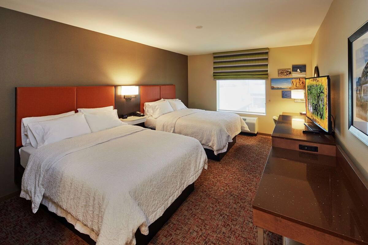 A hotel room with two beds, a desk, a television, and artwork on the wall.
