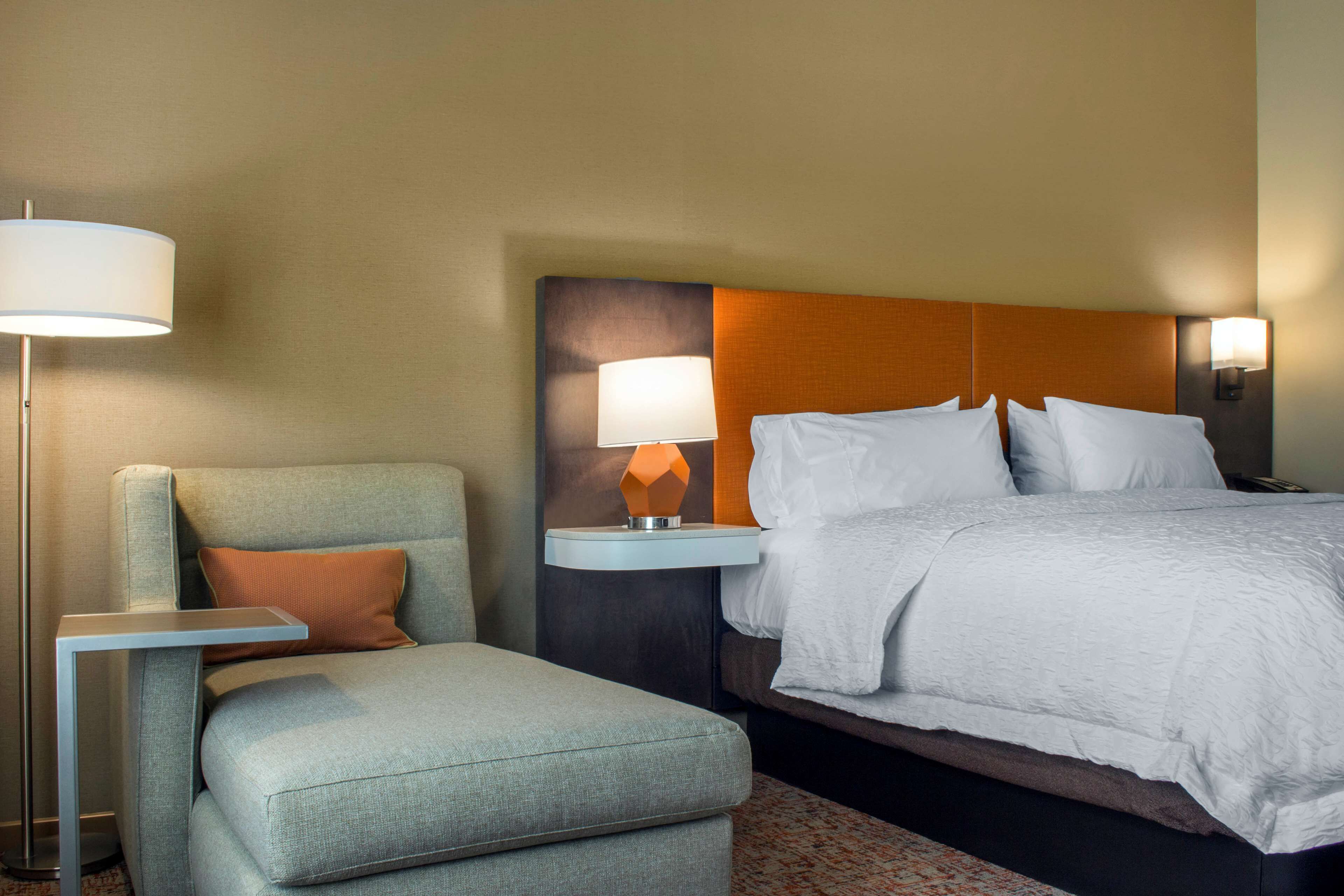 Quarto de hotel com cama, mesa de cabeceira com abajur, área de estar com poltrona e banquinho, e tapete estampado.