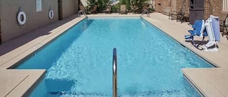 Una piscina rectangular con barandilla de seguridad metálica, rodeada de sillas de descanso y sombrillas.