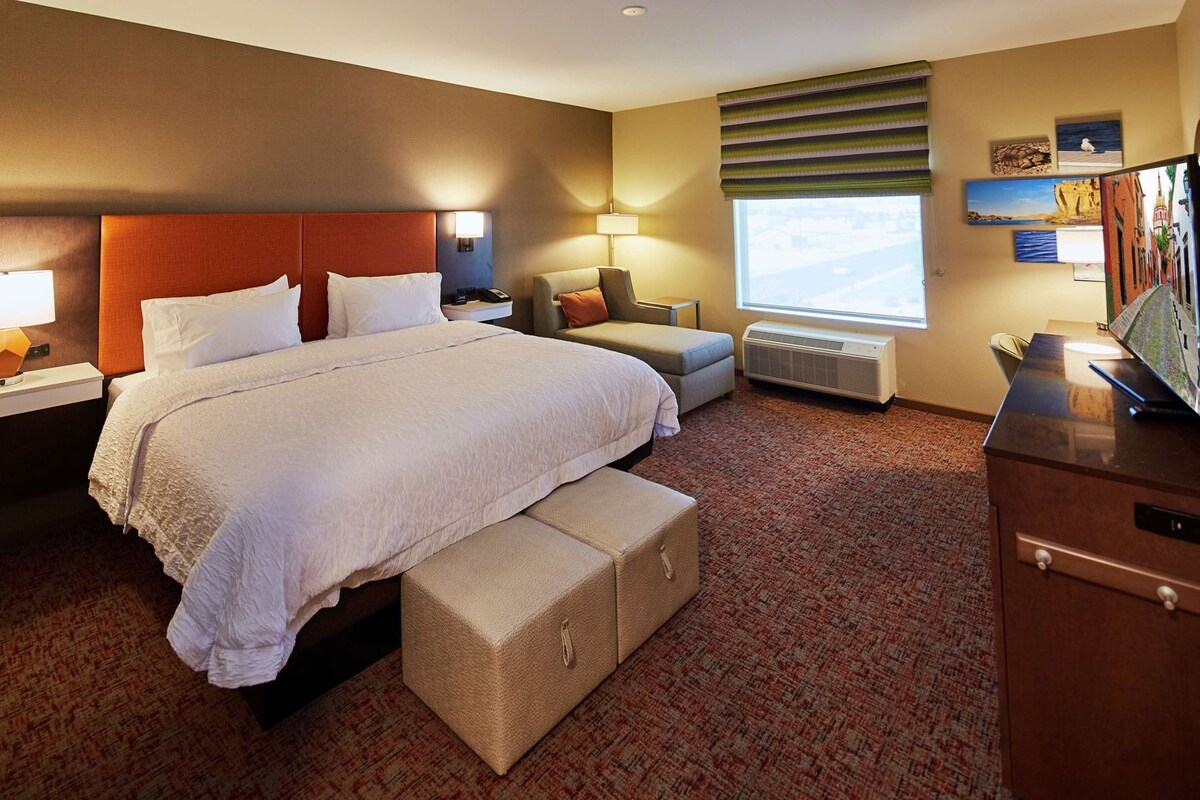 Habitación de hotel con una cama grande, un escritorio, una silla, un banco, un calentador y una ventana con vistas.