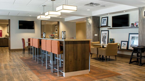 Un área de bar moderna con mostrador de madera y varios taburetes. Hay cuadros enmarcados en las paredes y un televisor montado sobre la barra.
