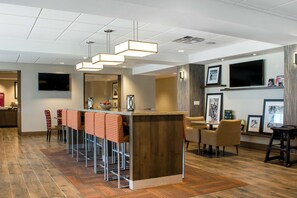 A modern bar area with a wooden bar counter and multiple stools. There are framed pictures on the walls and a television mounted above the bar.