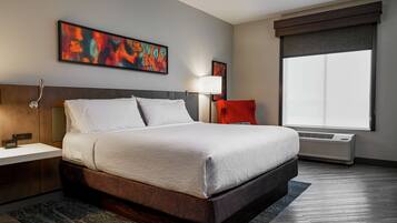 Quarto de hotel moderno com uma cama grande, uma mesa de cabeceira, uma luminária, uma cadeira vermelha e uma janela ampla.