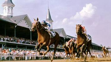 A horse race with jockeys on horses, a crowd of spectators, and a stadium with a distinctive architectural style.